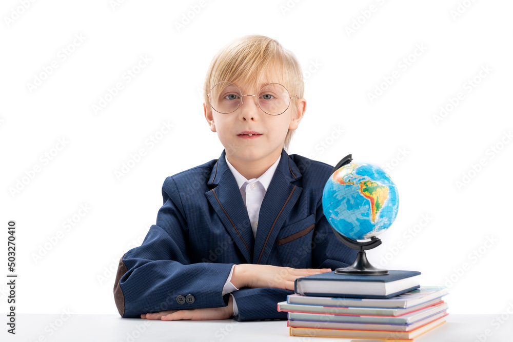Excellent boy sits at table with his hands folded and stares seriously at camera. First grader student next to notebooks and globe. Excellent boy sits at table with his hands folded and stares seriously at camera. First grader student next to notebooks and globe.