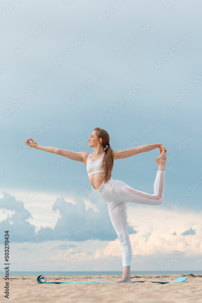 Fototapeta premium Young woman in white sportwear practicing yoga in the morning on seashore. Yoga at the beach. Doing asana Natarajasana
