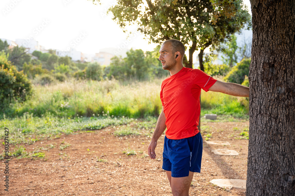 Caucasian man stretching after sport. He is leaning against a tree. He is wearing headphones, a red T-shirt and blue shorts. Spain. Caucasian man stretching after sport. He is leaning against a tree. He is wearing headphones, a red T-shirt and blue shorts. Spain.