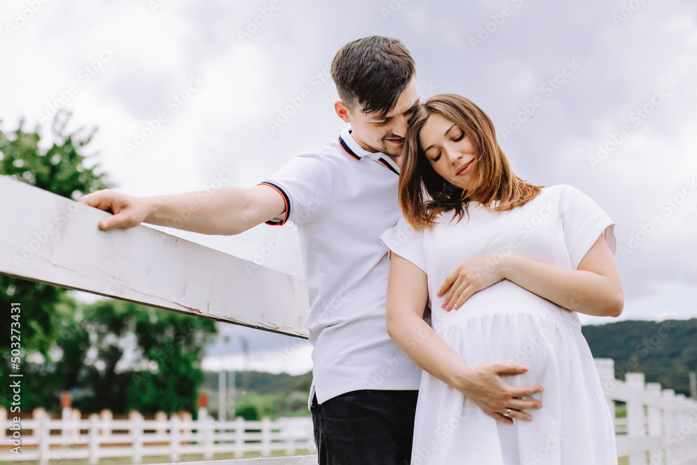 Naklejka premium A man hugs his pregnant wife near the white fence on a background of cloudy sky