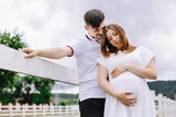 A man hugs his pregnant wife near the white fence on a background of cloudy sky