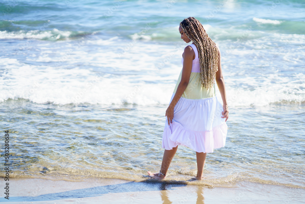 Black female walking along the shore of the beach wearing a beautiful long dress.