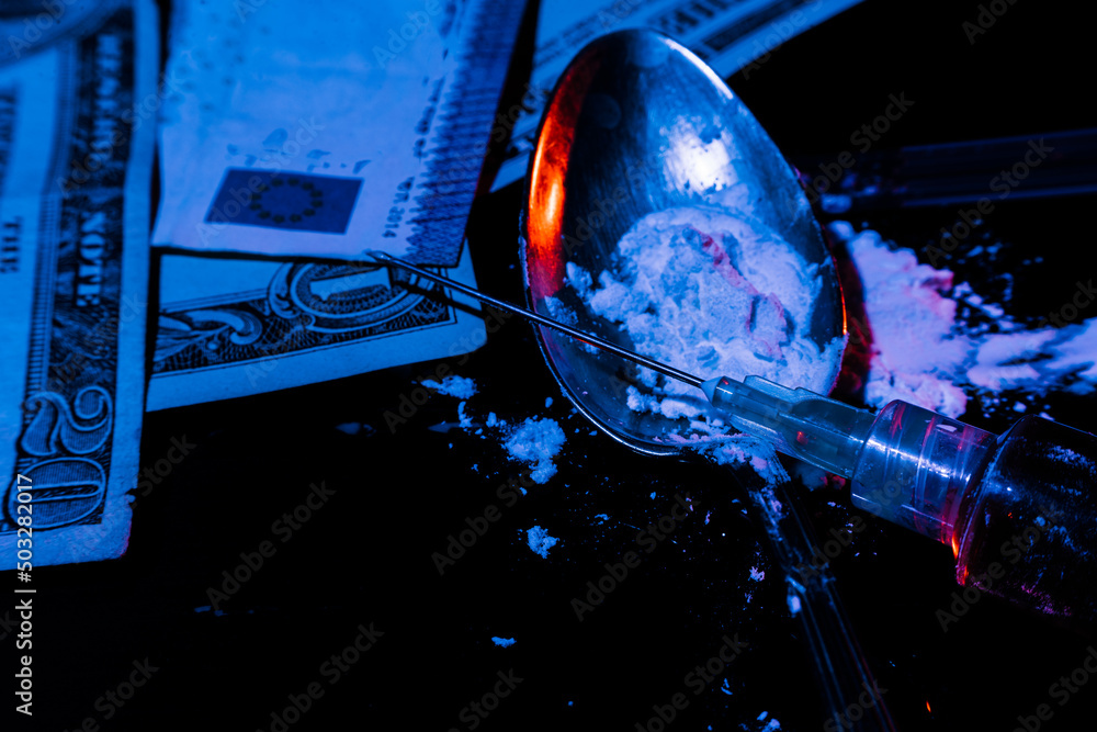 Syringe with cooked Heroin, white powder in a spoon and banknotes on a dark table with two-coloured lighting suggesting the forbidden and illegal. Concept of drug addiction, illegal trade. high view
