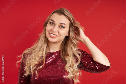 Beautiful blonde woman in a red dress on a red background touches her curls with her hand