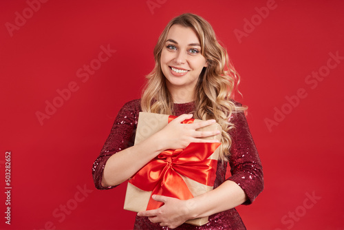 Beautiful cheerful blonde woman holds a gift box in her hands. Isolate on a red background