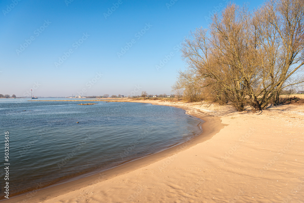 Sandy beach on the bank of the Dutch river Waal. It is a sunny day with a clear blue sky at the beginning of the spring season. River groynes made of basalt blocks are visible in the background.