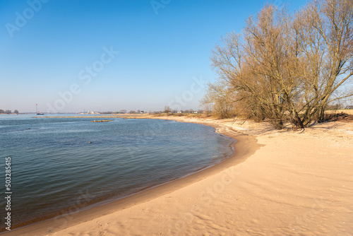 Fototapeta Naklejka Na Ścianę i Meble -  Sandy beach on the bank of the Dutch river Waal. It is a sunny day with a clear blue sky at the beginning of the spring season. River groynes made of basalt blocks are visible in the background.