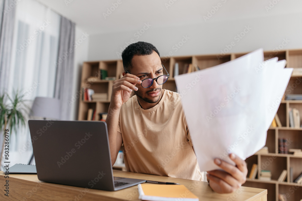 Serious young arab man reading documents and working on laptop remotely from home, sitting at desk Serious young arab man reading documents and working on laptop remotely from home, sitting at desk