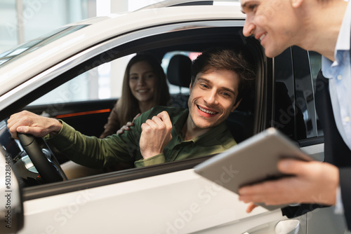 Young Caucasian couple clarifying car purchase details with salesman, looking at tablet screen in dealership center