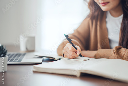 Close up - Hand of woman writing in spiral notepad placed on wooden desktop with various items at home. copy space.