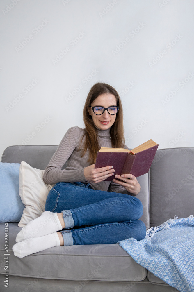 Smiling young woman in glasses sitting on sofa reading a book Smiling young woman in glasses sitting on sofa reading a book