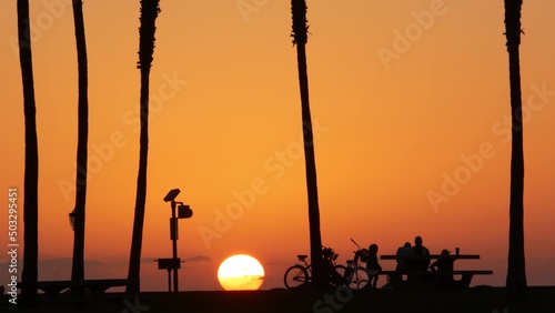 Orange sky  silhouettes of palm trees on beach at sunset  California coast  USA. Bicycle or bike in beachfront park at sundown in San Diego  Mission beach vacations resort on shore. People walking.