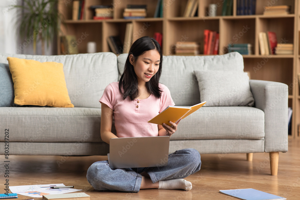 Young korean lady studying online from home, reading book and using laptop pc, sitting on floor near sofa, free space Young korean lady studying online from home, reading book and using laptop pc, sitting on floor near sofa, free space
