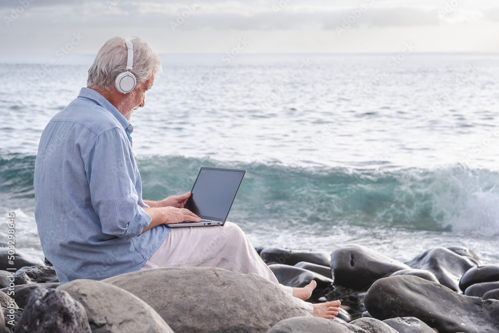 Senior active man sitting on the beach using laptop wearing headphones. Elderly bearded male barefoot enjoying sunset at sea, horizon over water