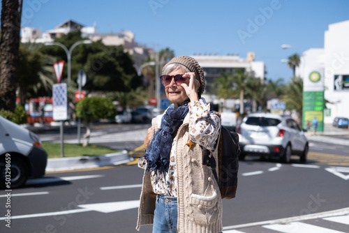 Smiling elderly woman traveler in sunny city center with backpack on shoulders while crossing the road on crosswalk.