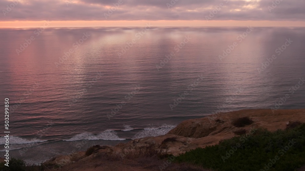 Dramatic cloudscape at sunset, reflection of pink sky, clouds. Torrey Pines scenic vista point, overlook viewpoint, ocean sea water waves from above. Steep cliff, rock or bluff, California coast, USA.