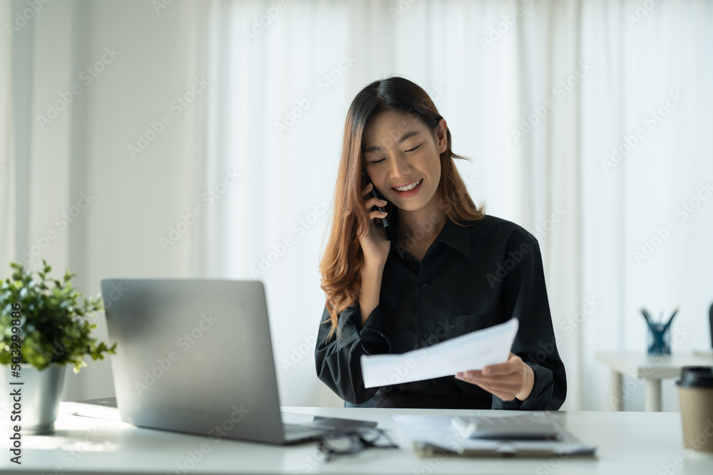 Young Asian Business Woman Using Computer in office with notebook and calling on mobile phone, financial accounting concept. Young Asian Business Woman Using Computer in office with notebook and calling on mobile phone, financial accounting concept.