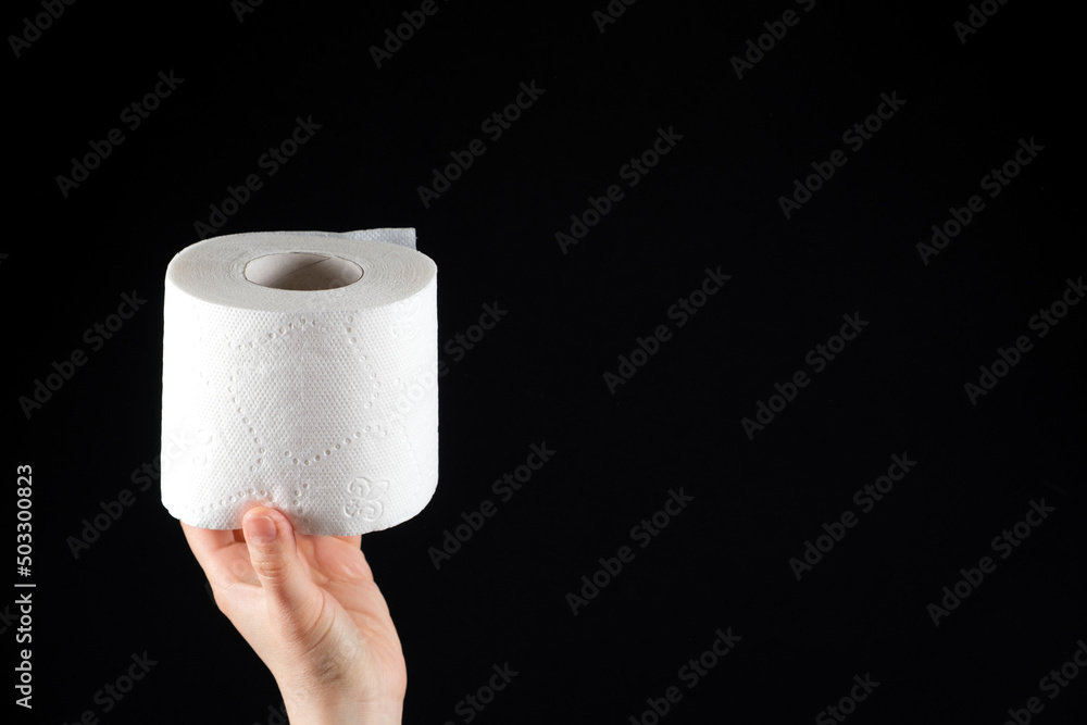 Fototapeta premium A roll of white soft toilet paper in a man's hand on a black background with space for text