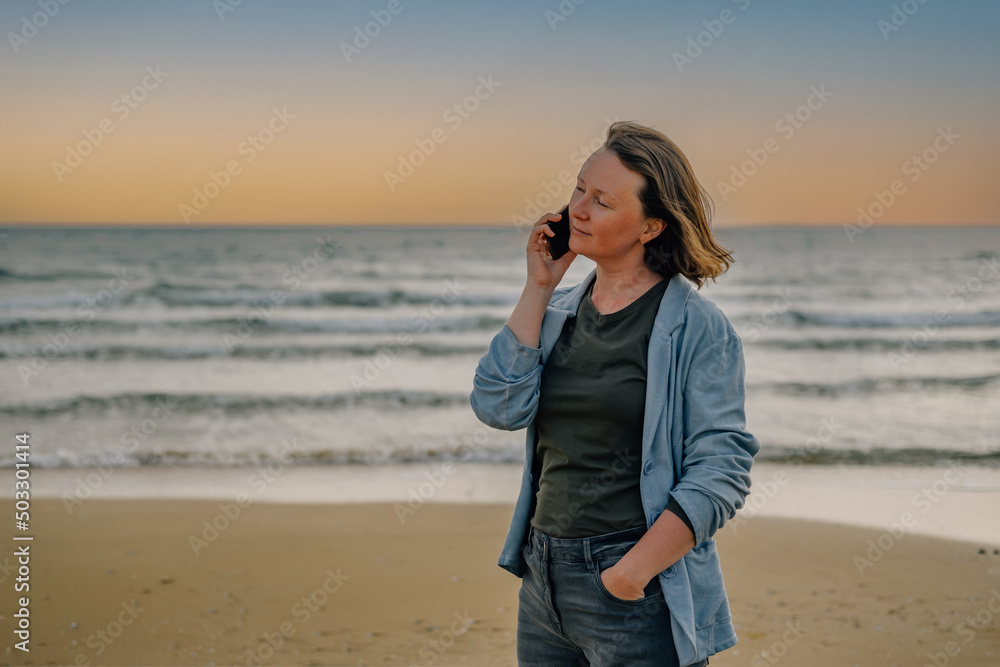 a young woman on the beach near the ocean in the spring at sunset talking on the phone with a smile on her face to her loved ones, friends to share the joy of vacation. modern technologies