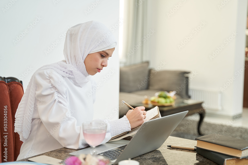 Muslim woman using laptop and writing in notebook Muslim woman using laptop and writing in notebook