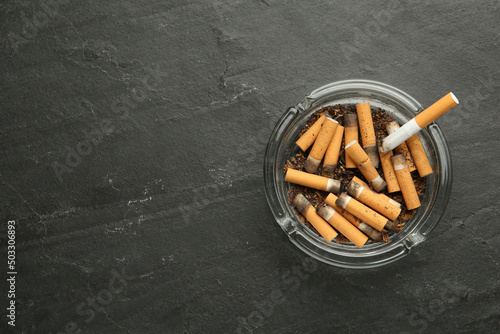 Glass ashtray with cigarette stubs on dark grey table, top view. Space for text