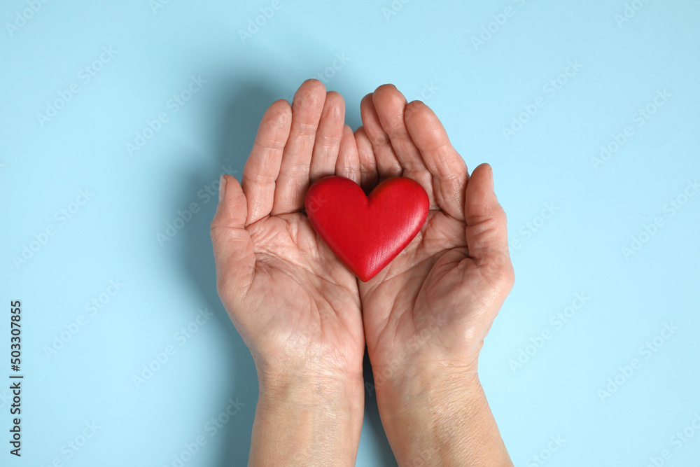 Elderly woman holding red heart in hands on light blue background, top view
