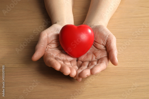 Elderly woman holding red heart in hands at wooden table  closeup