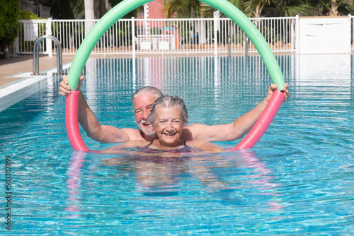 Fototapeta Naklejka Na Ścianę i Meble -  Active senior couple doing exercise in swimming pool with swim noodles. Happy retired people play in the outdoor pool water under the sun