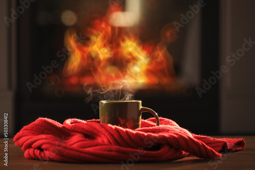 Cup of hot drink and red knitted scarf on wooden table near fireplace at home. Cozy atmosphere
