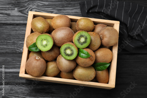 Crate with cut and whole fresh kiwis on black wooden table, top view