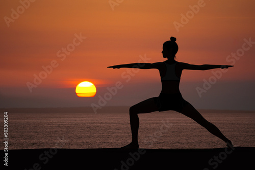 Woman silhouette in Yoga Warrior pose at sunset with quiet ocean