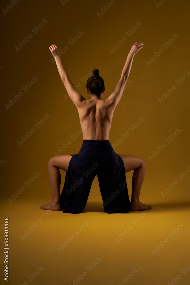 Obraz premium Back view of slim girl with hair bun standing with legs bent and hands raised. Young female with bare back stretching, yoga practicing. Isolated on yellow studio background.