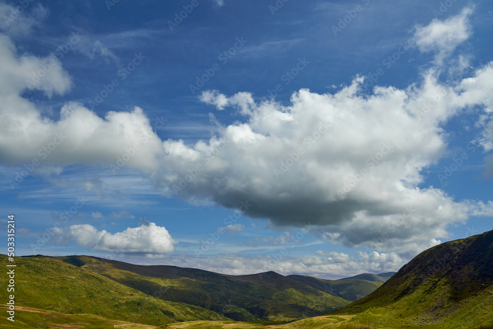 Huge cumulus clouds floating in blue sky, leaving shadows on grassy meadows outdoors. Panoramic view of sunlit valley between mountain hills in sunny day, with copy space. Concept of landscape. 