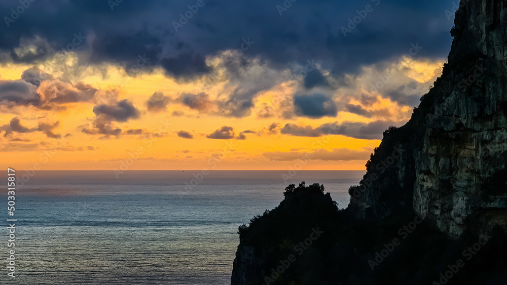 Viewing the sunset next to a sharp cliff at the Mediterranean sea at Amalfi Coast, Campania, Italy, Europe. Golden hour sunlight on surface of Tyrrhenian Sea. Red orange clouds emerging. Coastline Viewing the sunset next to a sharp cliff at the Mediterranean sea at Amalfi Coast, Campania, Italy, Europe. Golden hour sunlight on surface of Tyrrhenian Sea. Red orange clouds emerging. Coastline