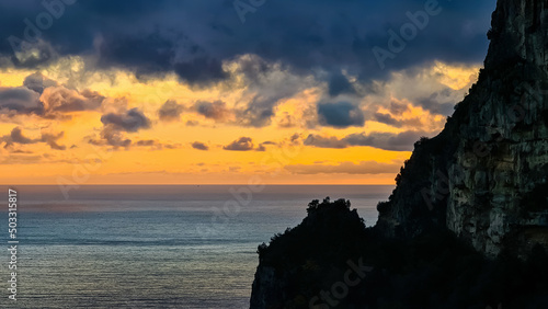 Viewing the sunset next to a sharp cliff at the Mediterranean sea at Amalfi Coast, Campania, Italy, Europe. Golden hour sunlight on surface of Tyrrhenian Sea. Red orange clouds emerging. Coastline