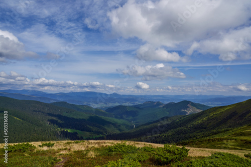 Scenic view from slope on covered with dry grass meadow between mountain peaks in early autumn. Panoramic view of woodland hills, with spectacular mountain range on background. Concept of landscape.