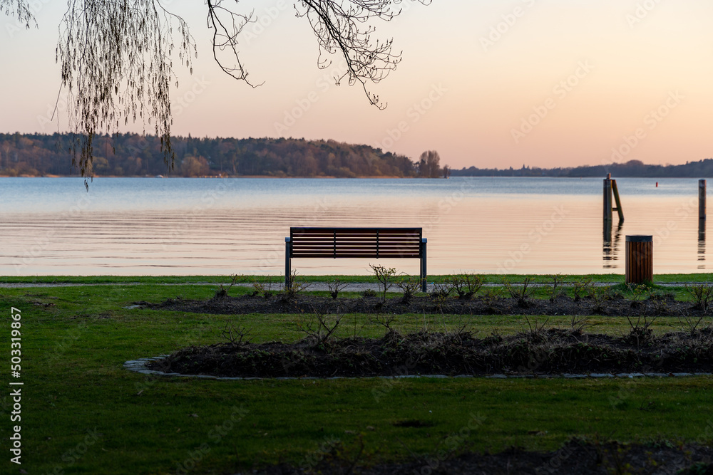 Empty park bench in front of a calm lake landscape. Place to relax in a sunset scene. Public park during spring season. Fresh green grass in the evening light. Travel destination in Germany.