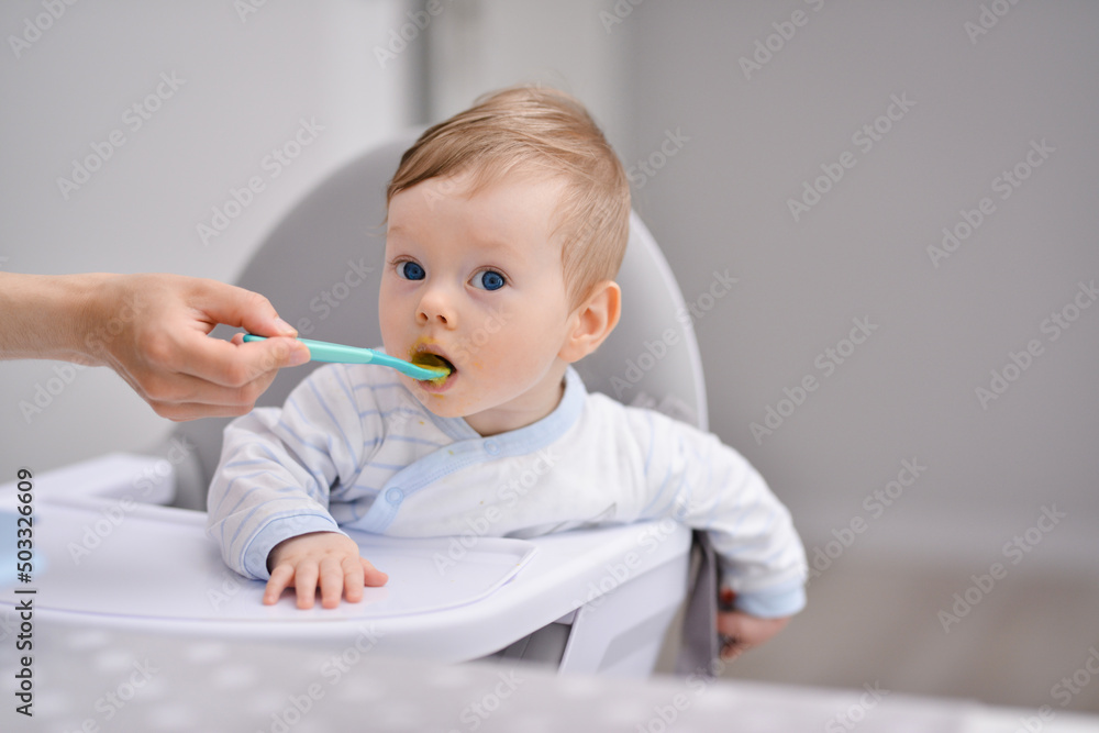 Mother feeds baby boy with a spoon porridge. Portrait of happy kid boy in high chair. Small child sits on a chair and eating with spoon. Mother feeds baby boy with a spoon porridge. Portrait of happy kid boy in high chair. Small child sits on a chair and eating with spoon.