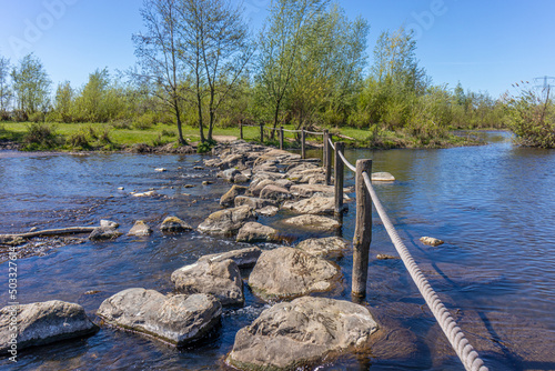 Oude Maas river with stepping stones used as a bridge at Brug Molenplas, wooden posts and rope fences, green trees in the background, sunny day in Stevensweert, South Limburg, Netherlands photo