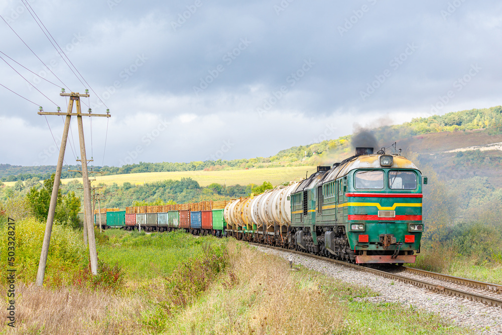A green locomotive pulls a train loaded with gasoline and timber to the railway station. Carpathian mountains. Trees with green and yellow leaves. Evening autumn lighting. A green locomotive pulls a train loaded with gasoline and timber to the railway station. Carpathian mountains. Trees with green and yellow leaves. Evening autumn lighting.