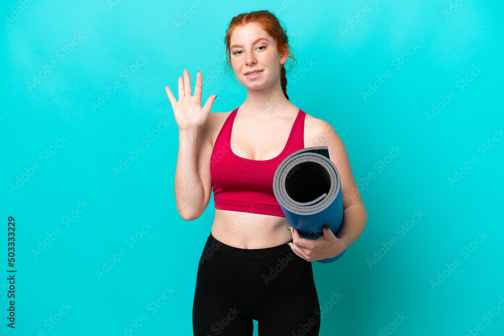 Young sport reddish woman going to yoga classes while holding a mat isolated on blue background saluting with hand with happy expression Young sport reddish woman going to yoga classes while holding a mat isolated on blue background saluting with hand with happy expression