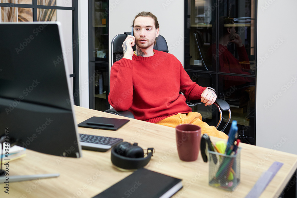 Young businessman sitting on chair in front of his workplace with computer and talking on mobile phone Young businessman sitting on chair in front of his workplace with computer and talking on mobile phone