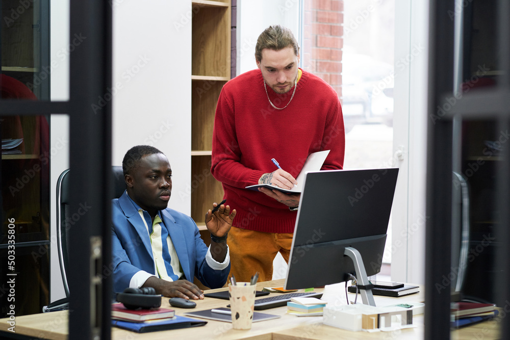 African manager sitting at his workplace with computer and giving work to businessman while he making notes in his notebook African manager sitting at his workplace with computer and giving work to businessman while he making notes in his notebook