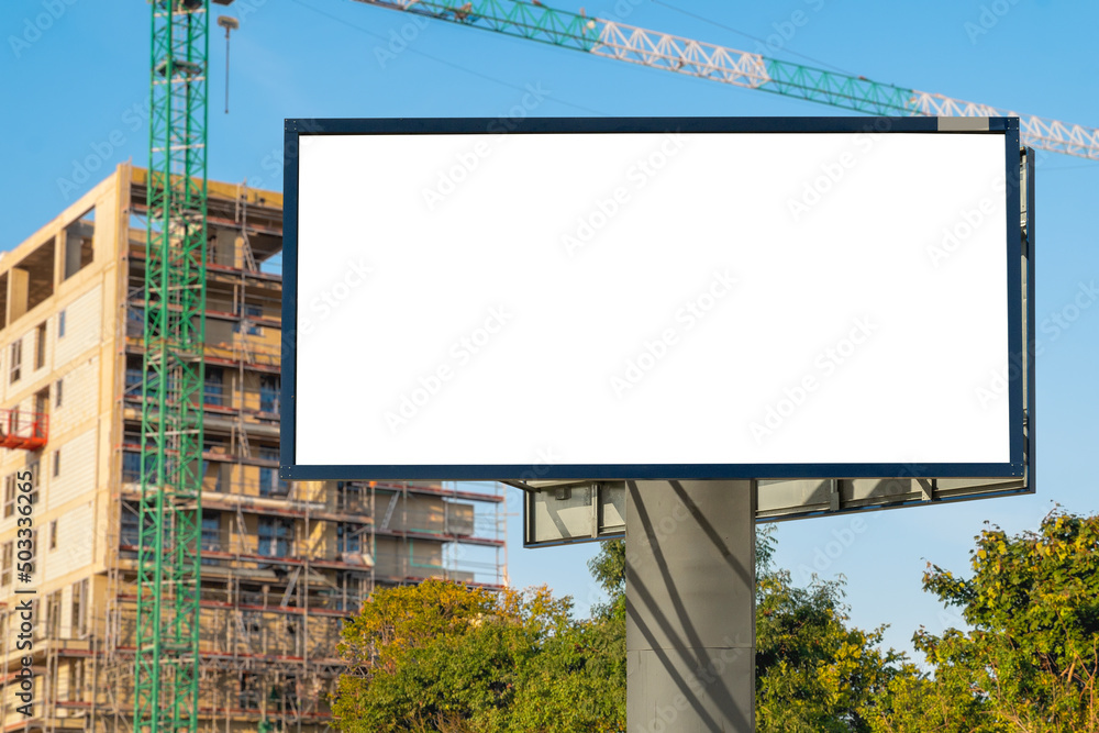 Blank white billboard for advertisement in front of the construction site. There is modern apartment building in the background. Blank white billboard for advertisement in front of the construction site. There is modern apartment building in the background.