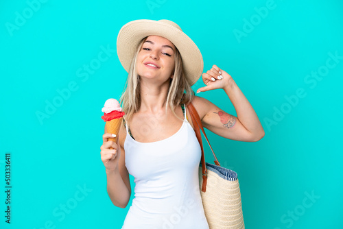 Young Romanian woman holding ice cream and beach bag isolated on blue background proud and self-satisfied