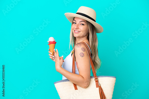 Young Romanian woman holding ice cream and beach bag isolated on blue background and pointing it