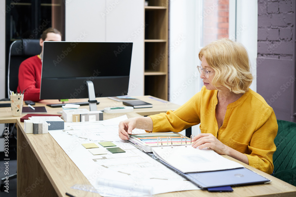 Designer deciding colour samples at table with her colleague working on computer in background Designer deciding colour samples at table with her colleague working on computer in background
