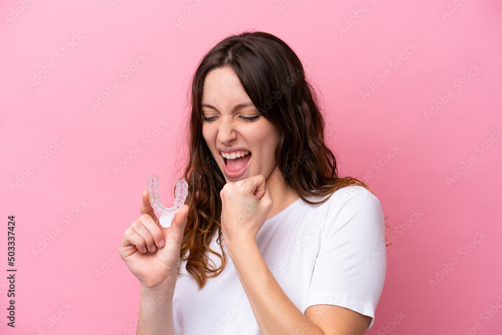 Young caucasian woman holding invisible braces isolated on pink background celebrating a victory Young caucasian woman holding invisible braces isolated on pink background celebrating a victory