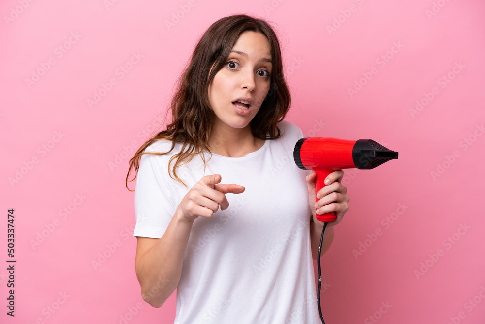 Young caucasian woman holding a hairdryer isolated on pink background surprised and pointing front Young caucasian woman holding a hairdryer isolated on pink background surprised and pointing front