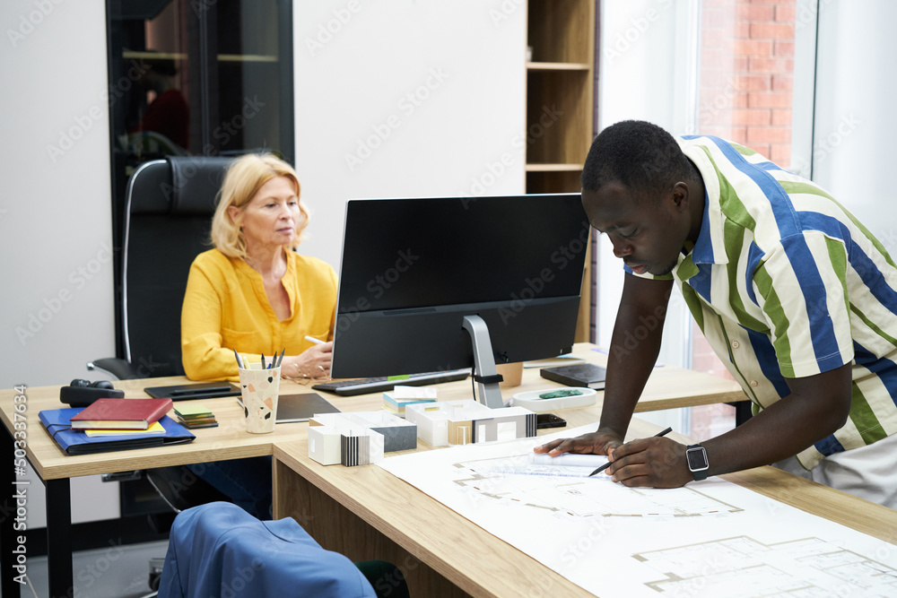 African male engineer drawing blueprint at table with his colleague working on computer in background African male engineer drawing blueprint at table with his colleague working on computer in background